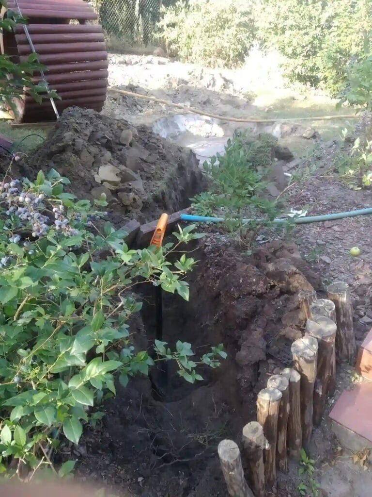 Precyzyjny, głęboki wykop ręczny (poniżej strefy mrozu) prowadzony w zadbanym ogrodzie, z chirurgicznym ominięciem krzewów borówki i palisady.