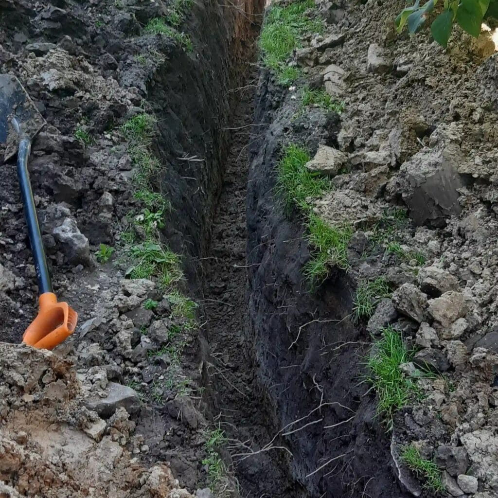 Precyzyjny, głęboki wykop pod przyłącze wodne w ciężkim gruncie, z darnią trawnika starannie odłożoną na bok w celu ochrony ogrodu.