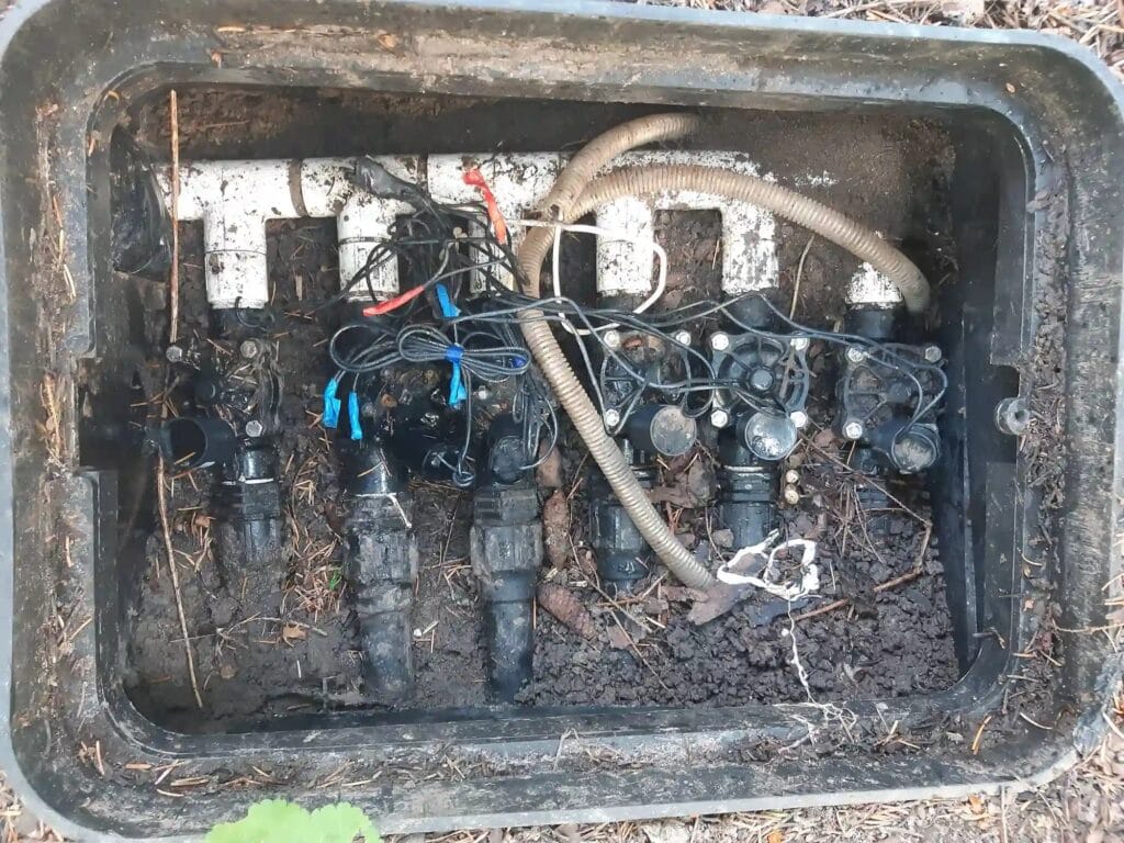 Patologiczny stan skrzynki zaworowej: elektrozawory zasypane błotem i piachem, plątanina kabli i brak możliwości serwisu.