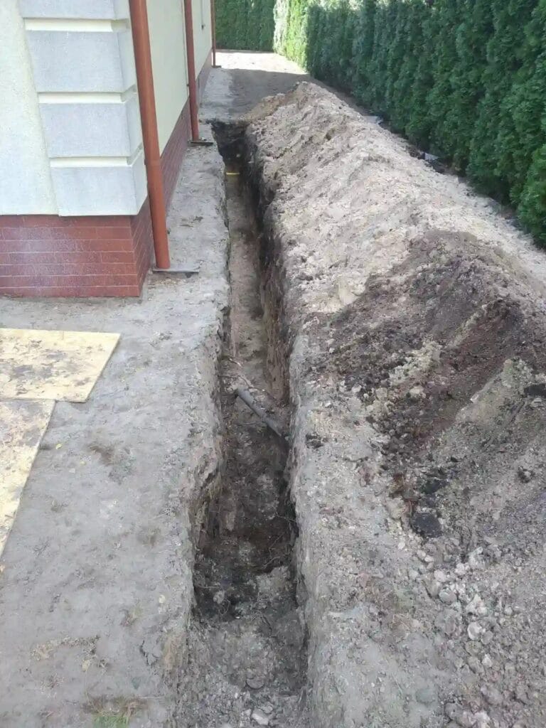 Głęboki, precyzyjny wykop liniowy (tranzyt) wzdłuż fundamentu budynku, przygotowany pod ułożenie rury PE poniżej strefy przemarzania.