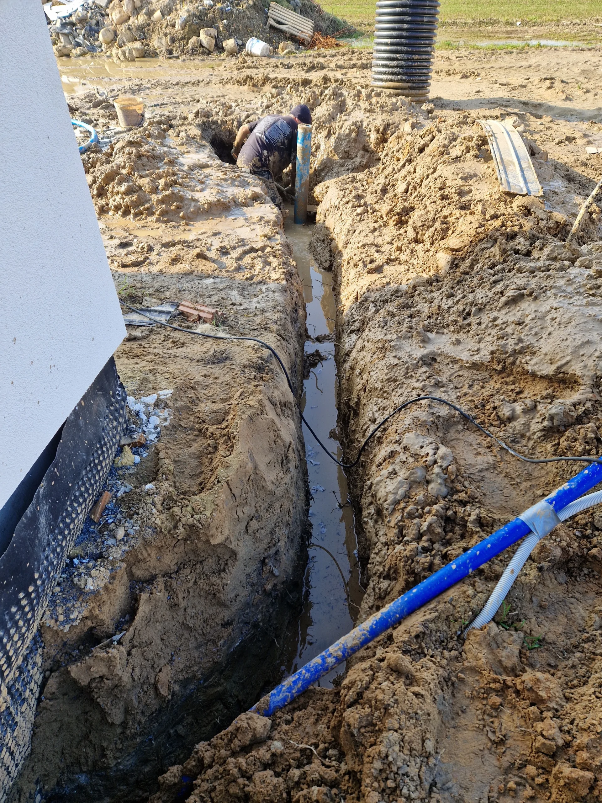Ciężka praca Hydromonters.pl: instalacja rury PE w głębokim, podmokłym wykopie, z widocznym drenażem (niebieska rura).