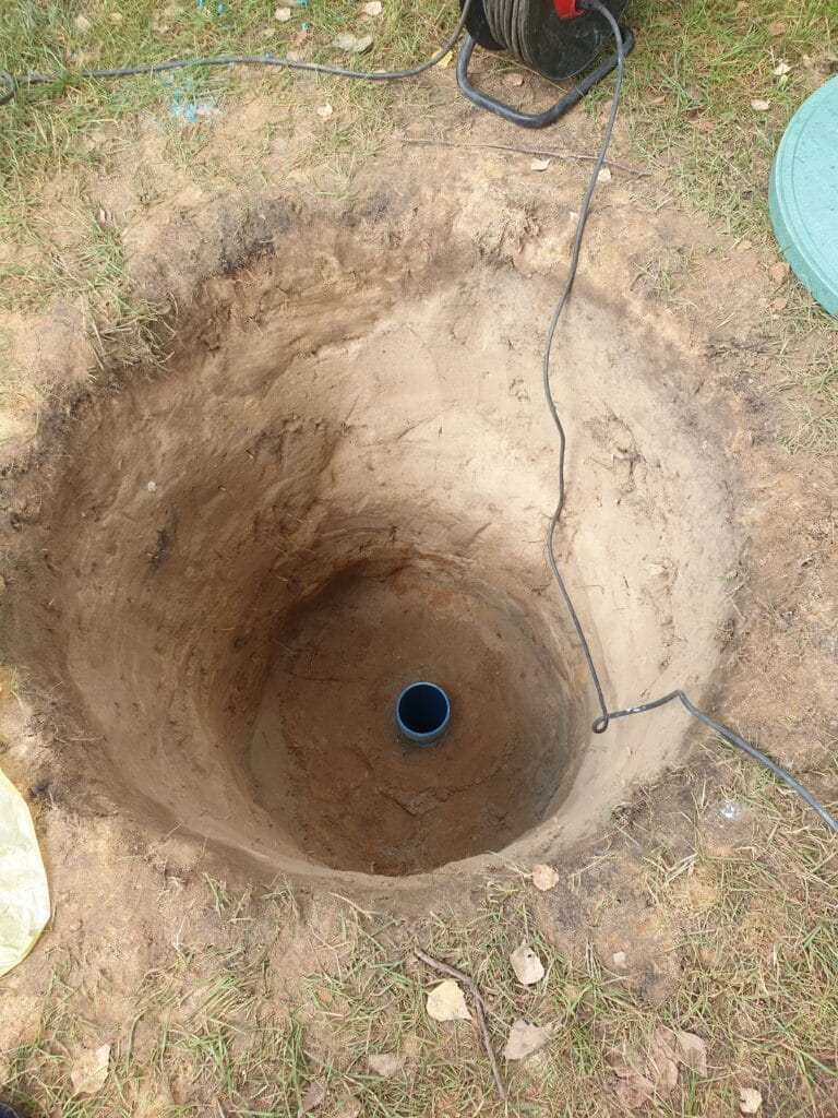 Odkopana rura osłonowa (niebieska) studni głębinowej na dnie szerokiego wykopu, przygotowana do montażu obudowy (komory rewizyjnej).