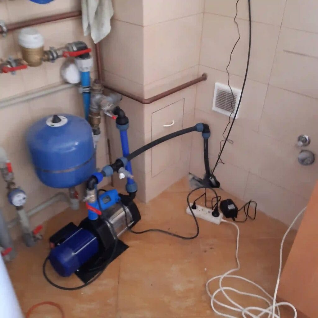 Niebezpieczna instalacja hydroforowa w piwnicy z pompą powierzchniową i przedłużaczem na podłodze, stwarzająca ryzyko porażenia prądem.