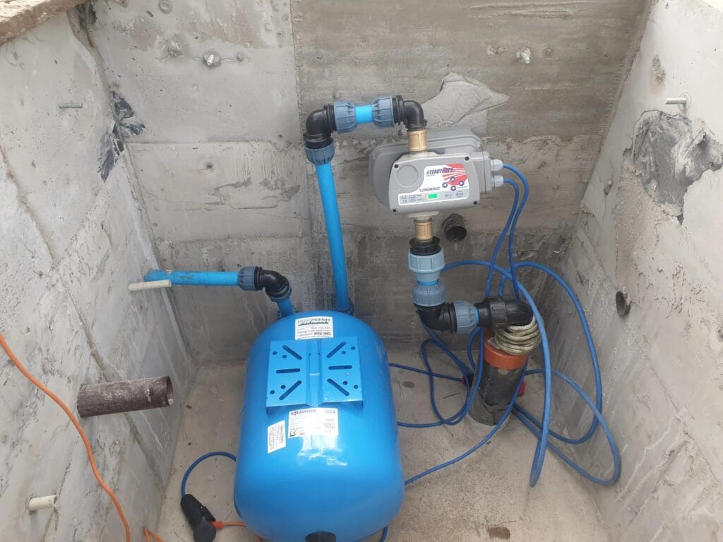 Standardowy montaż nowoczesnego zestawu hydroforowego w betonowej komorze rewizyjnej. Widoczny niebieski zbiornik przeponowy (membranowy), pompa oraz automatyczny sterownik ciśnieniowy (automat) zapewniający bezobsługową pracę.