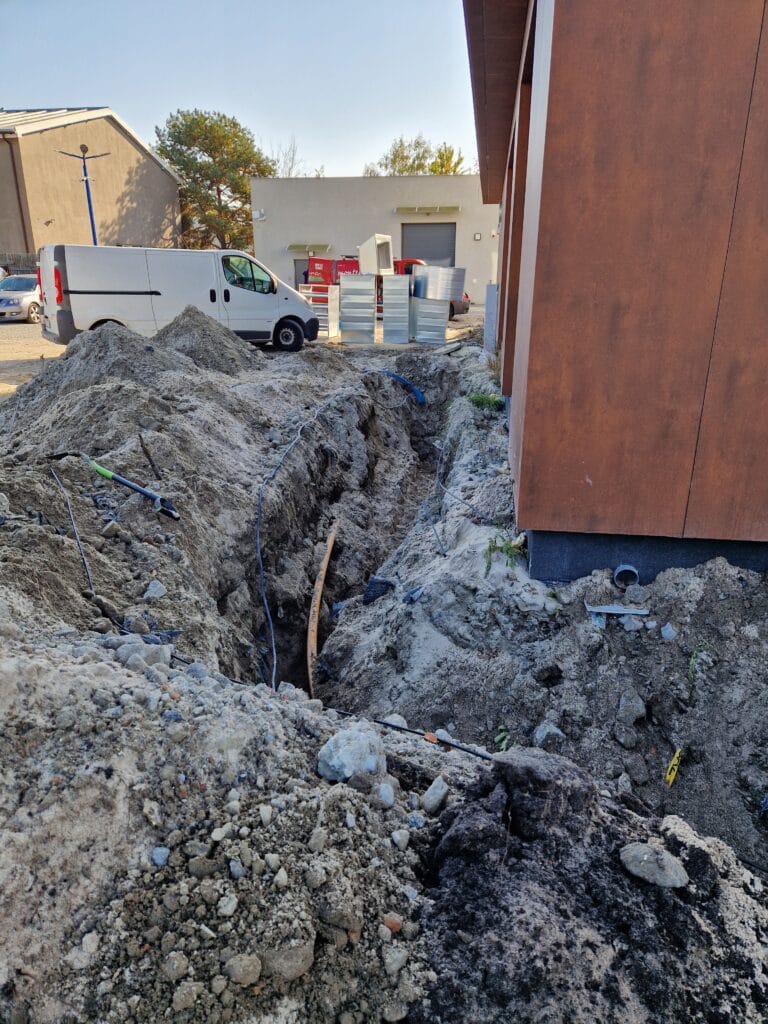 Ekstremalnie trudne prace ziemne Hydromonters.pl przy nowym budynku. Głęboki wykop (poniżej strefy przemarzania) na rurę PE i kabel, w tle auto serwisowe.