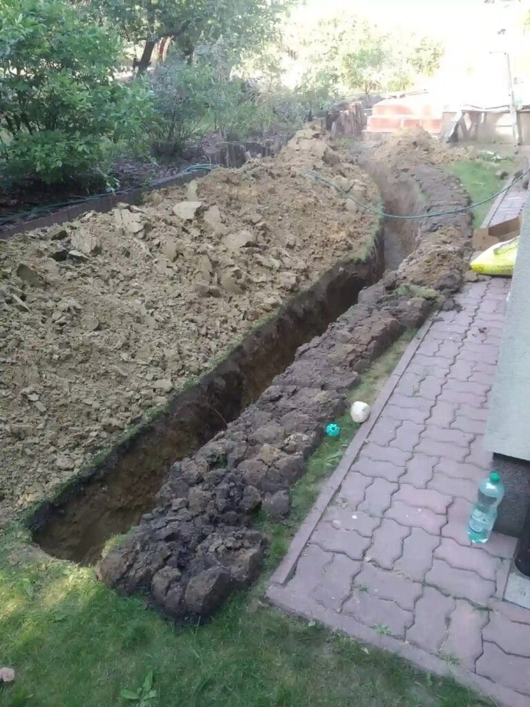 Tytaniczna praca Hydromonters.pl: głęboki, długi wykop pod przyłącze wodne, z urobkiem składowanym na plandece i starannie odłożoną darnią.