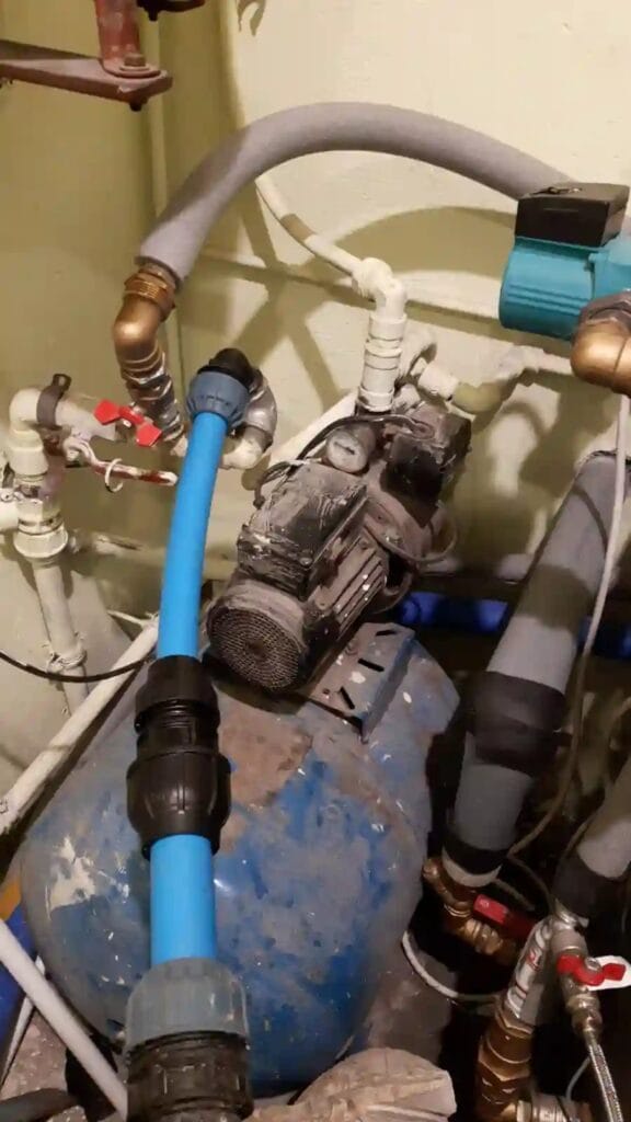 Proces modernizacji starej, skomplikowanej instalacji hydroforowej w warszawskiej kotłowni. Widoczna stara pompa, zbiornik oraz nowe, niebieskie rury PE wprowadzane do systemu.