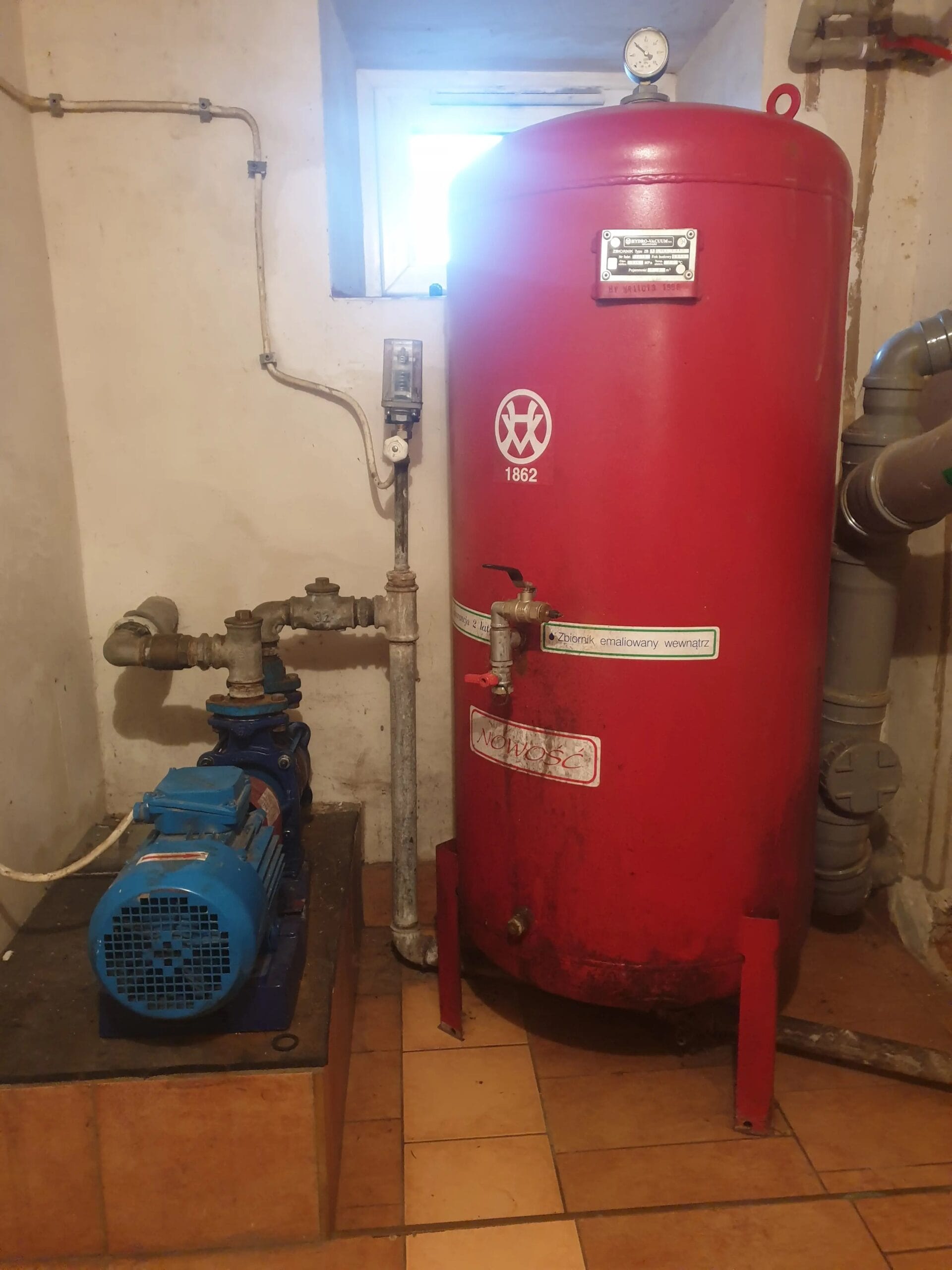 Serwis dużej instalacji hydroforowej w kotłowni. Widoczna pompa Hydro-Vacuum S.A. oraz duży, pionowy zbiornik hydroforowy z manometrem.