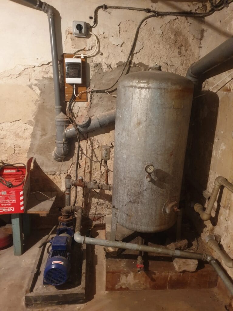 Ewidentne zagrożenie: stary zbiornik ocynkowany i pompa hydroforowa z instalacją elektryczną na wilgotnej, zagrzybionej ścianie piwnicy.