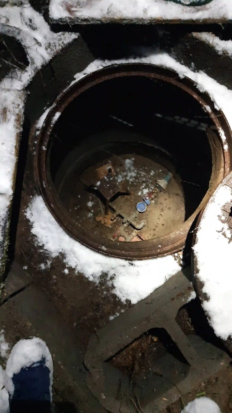 Awaryjny serwis pompy głębinowej w środku zimy. Serwisant przy ośnieżonym włazie do studni w nocy.