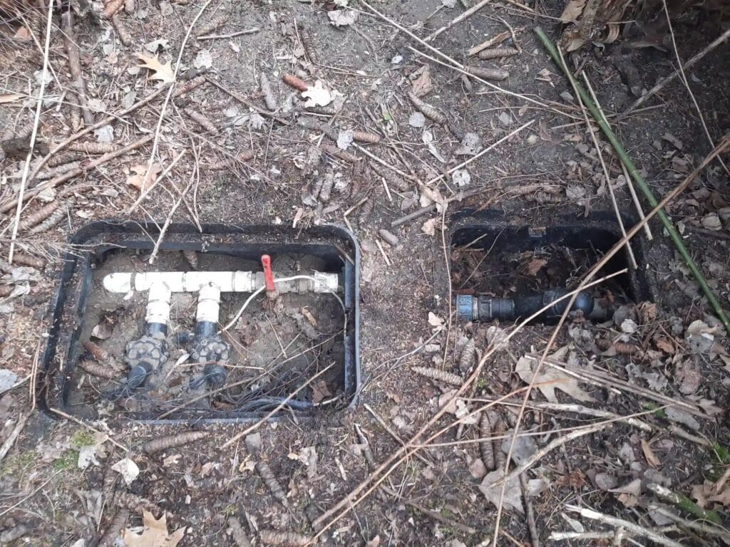 Zaniedbana, otwarta skrzynka rozdzielacza automatycznego nawadniania z elektrozaworami zalanymi piaskiem, gałęziami i zanieczyszczeniami. Obraz ilustrujący potrzebę regularnej konserwacji.