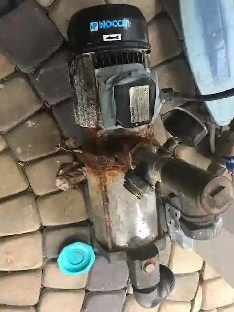 Zardzewiały i uszkodzony korpus pompy hydroforowej NOCCHI leżącej na kostce brukowej. Widoczny wyciek i ślady korozji świadczące o krytycznej awarii i konieczności wymiany na nowy model.