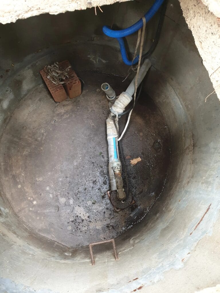 Stara i zaniedbana instalacja pompy w betonowej studni kręgowej, z widocznymi amatorskimi łączeniami rur, prowizorycznymi mocowaniami i zanieczyszczeniami. Obraz ilustrujący potrzebę modernizacji.