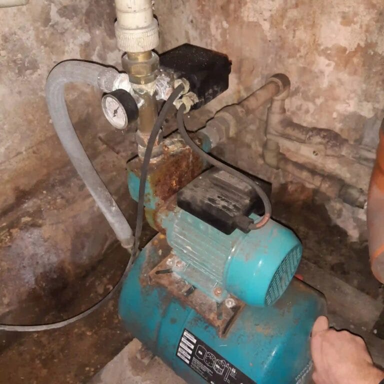 Stary, skorodowany zestaw hydroforowy (pompa WZ-750) w wilgotnej piwnicy, z krytycznym błędem w postaci elastycznego węża na ssaniu.