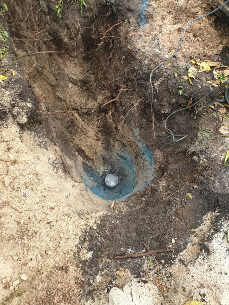 Montaż hermetycznej głowicy studziennej (srebrna) na niebieskiej rurze osłonowej, na dnie głębokiego wykopu roboczego.