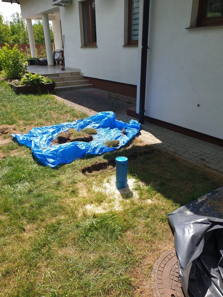 Zakończony odwiert studni głębinowej obok domu; widoczna niebieska rura osłonowa i plandeka z darnią, chroniąca trawnik przed zniszczeniem.