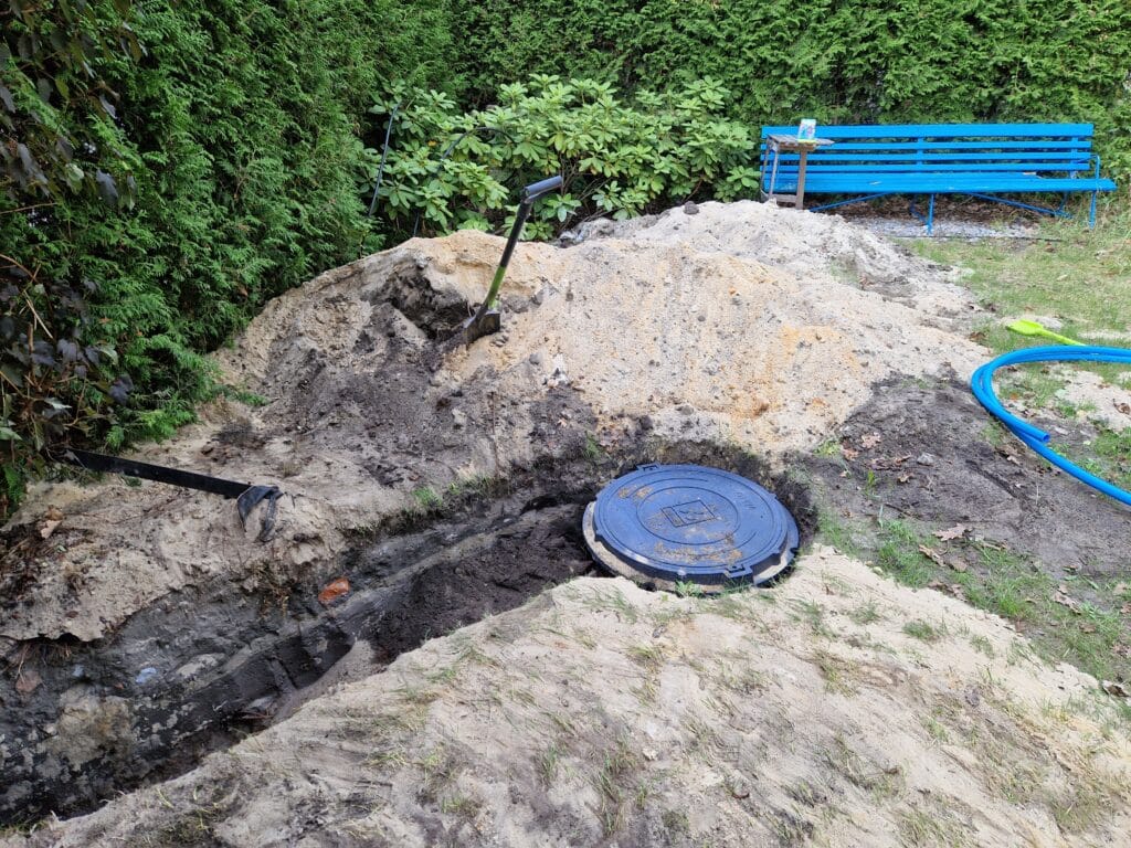 Zwieńczenie prac Hydromonters.pl: obudowa studni głębinowej (sucha komora rewizyjna) zakończona trwałym włazem, obok głębokiego wykopu pod przyłącze.