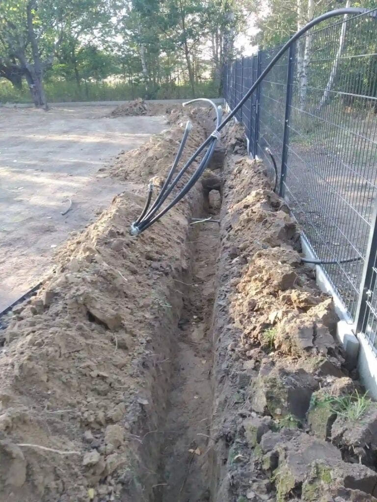 Montaż magistrali automatycznego systemu nawadniania: grube rury PE ułożone w wykopie wzdłuż ogrodzenia, zasilające przyszłe elektrozawory i zraszacze.