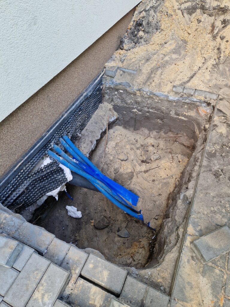 Przemyślane wprowadzenie przyłącza wodnego do budynku: rury PE i kable elektryczne w peszlach ochronnych (rurach osłonowych) w wykopie przy fundamencie.