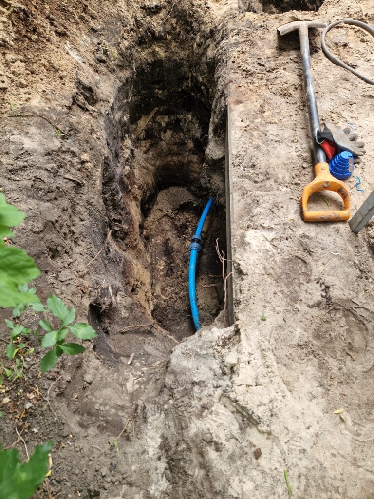 Ziemne prace Hydromonters.pl: głęboki wykop i bezpieczne wyprowadzenie niebieskiej rury PE z obudowy studni głębinowej.