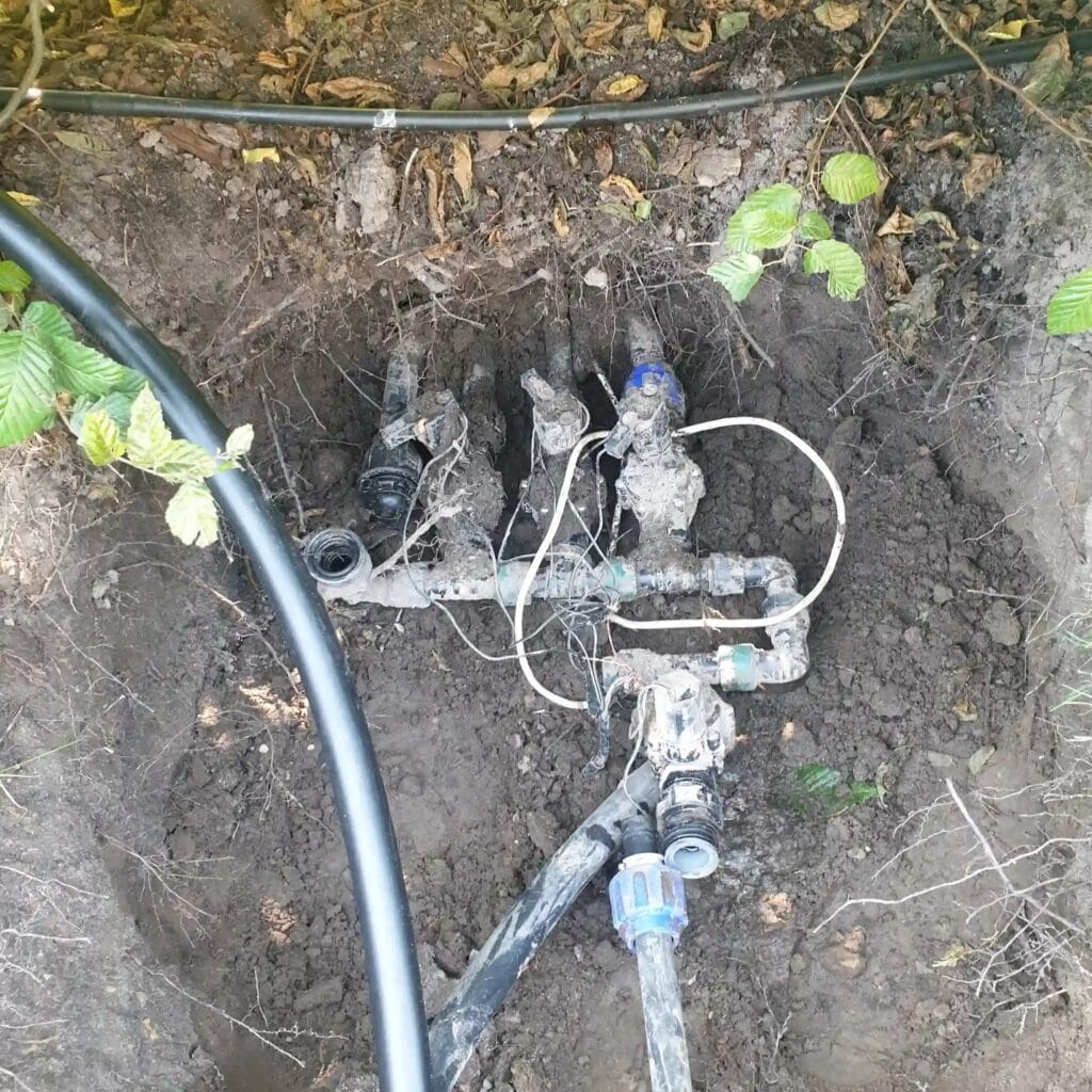 Serwis i wymiana elektrozaworów w sekcjach automatycznego systemu nawadniania ogrodu. Odsłonięta, podziemna skrzynka z kolektorem i przewodami sterującymi, wymagająca naprawy.