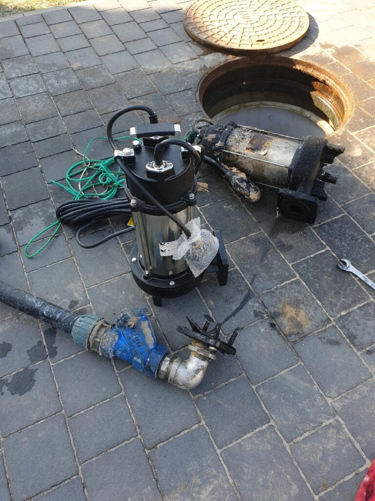 Wymiana uszkodzonej pompy zatapialnej do ścieków na nowy, wysokiej jakości model z wirnikiem rozdrabniającym. Pompy leżą na kostce brukowej obok otwartego włazu rewizyjnego szamba.